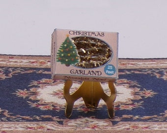 Könnte beinhalten: Eine Miniaturbox mit goldener Weihnachtsgirlande, 7,6 Meter lang, mit einem kleinen Weihnachtsbaum auf der Vorderseite der Box. Die Box steht auf einem goldenen Ständer.