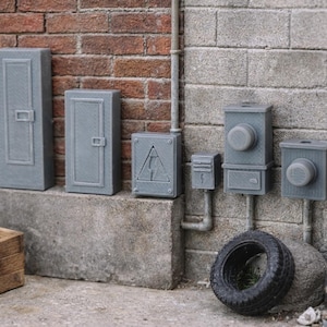 May include: Miniature gray electrical boxes and panels are mounted on a brick and concrete wall. A tire, wooden crate, and metal barrel are in the foreground. The electrical panel has a high voltage warning symbol.