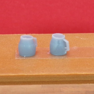 May include: Two miniature light blue mugs with rounded bodies and small side handles. These small vessels feature a smooth, solid-colored finish and an open top design, presented as a pair of identical items for display or collection purposes.
