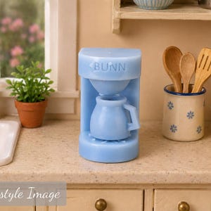 May include: A light blue miniature coffee maker with the word BUNN embossed on the front. The device features a cylindrical base, a central brewing area, and a small matching light blue carafe with a handle, designed for a dollhouse kitchen setting.