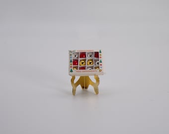 Könnte beinhalten: Eine Schachtel mit Miniatur-Weihnachtsbaumschmuck in Rot, Gold und Silber, präsentiert auf einem goldfarbenen Ständer. Auf der Schachtel steht "Christmas Tree Ornaments". Die Kugeln sind klein, rund und glänzend.