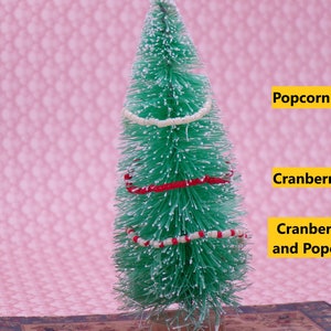 May include: A small artificial Christmas tree decorated with white, red, and white and red popcorn garlands. The tree is on a pink background with the words "Popcorn", "Cranberries", and "Cranberries and Popcorn" written in yellow.