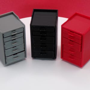 May include: Three miniature storage cabinets in gray, black, and red. Each cabinet has five drawers and a flat top. The cabinets are arranged side by side on a white surface, with a red background.