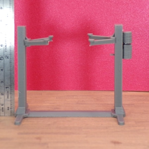 K&ouml;nnte beinhalten: Graues Modell einer Hebeb&uuml;hne, ca. 13 cm hoch, mit Sockel und zwei vertikalen St&uuml;tzen. Die Hebeb&uuml;hne hat Arme, die sich von oben erstrecken, um ein Fahrzeug zu halten. Ein Metalllineal liegt daneben, vor rotem Hintergrund.