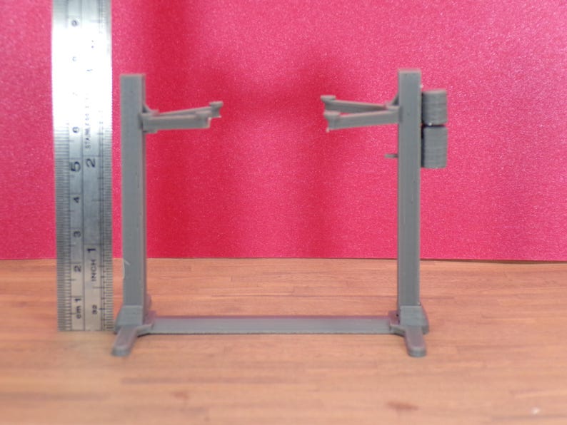 K&ouml;nnte beinhalten: Graues Modell einer Hebeb&uuml;hne, ca. 13 cm hoch, mit Sockel und zwei vertikalen St&uuml;tzen. Die Hebeb&uuml;hne hat Arme, die sich von oben erstrecken, um ein Fahrzeug zu halten. Ein Metalllineal liegt daneben, vor rotem Hintergrund.