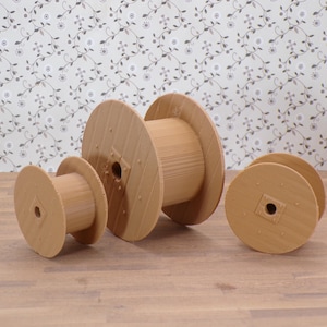 May include: Four wooden spools of varying sizes are arranged on a wooden surface. The spools are light brown and have a cylindrical shape with a central hole. The background features a floral patterned wallpaper.