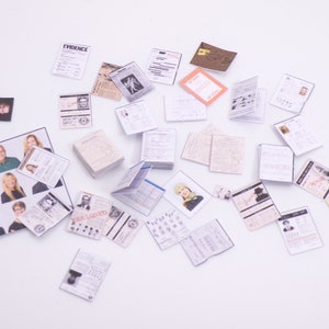 Peut inclure: Une collection de documents papier miniatures, dont une pile de papiers &eacute;tiquet&eacute;s "EVIDENCE", un article de journal et plusieurs cartes d'identit&eacute; avec des photos de personnes. Les documents sont &eacute;parpill&eacute;s sur une surface blanche.