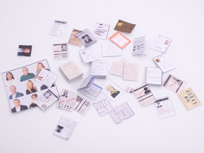 Peut inclure: Une collection de documents papier miniatures, dont une pile de papiers &eacute;tiquet&eacute;s "EVIDENCE", un article de journal et plusieurs cartes d'identit&eacute; avec des photos de personnes. Les documents sont &eacute;parpill&eacute;s sur une surface blanche.