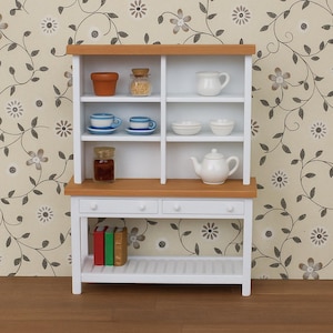 May include: A miniature white and wood-toned kitchen hutch with various small kitchen items. The hutch has shelves holding a small terracotta pot, a jar with a cork lid, a white pitcher, two blue and white teacups, small white bowls, a jar of preserves, and a white teapot.