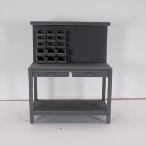 May include: A gray miniature workbench with a backboard and storage bins. The workbench has two drawers and a lower shelf. The backboard features a series of small storage compartments. The workbench is made of a solid, matte material.