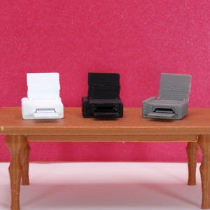 May include: Three miniature printers in white, black, and gray sit on a small brown table against a pink background. The printers have a rectangular design with a paper output slot and a raised top.