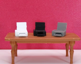 May include: Three miniature printers in white, black, and gray sit on a small brown table against a pink background. The printers have a rectangular design with a paper output slot and a raised top.