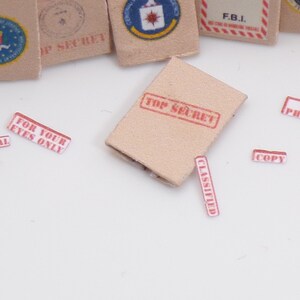 Peut inclure: Une enveloppe en papier brun avec un tampon rouge "TOP SECRET". Il y a plusieurs autres autocollants rouges et blancs avec du texte comme "CONFIDENTIEL", "NE PAS PHOTOCOPIER", "POUR VOS YEUX SEULEMENT", "CLASSIFIE", "COPIE", "SECURISE", et "TOP SECRET".
