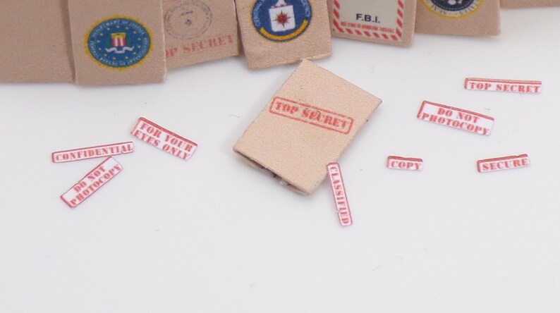 Peut inclure: Une enveloppe en papier brun avec un tampon rouge "TOP SECRET". Il y a plusieurs autres autocollants rouges et blancs avec du texte comme "CONFIDENTIEL", "NE PAS PHOTOCOPIER", "POUR VOS YEUX SEULEMENT", "CLASSIFIE", "COPIE", "SECURISE", et "TOP SECRET".