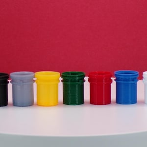 May include: Seven small, cylindrical containers in a row, each in a different colour: black, grey, yellow, green, red, blue, and white. They have ridged exteriors and wider rims. The background is a solid red.