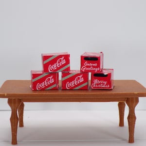 May include: Miniature Coca-Cola boxes in red and white with diagonal stripes and the Coca-Cola logo. Some boxes have holiday greetings. The boxes are arranged on a small brown table with curved legs.