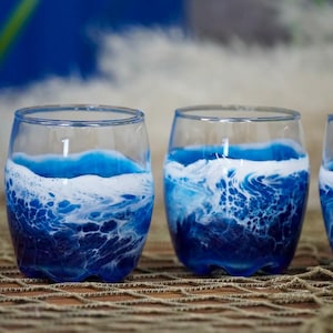 May include: Clear stemless glasses with a blue and white wave design. The blue resin mimics ocean waves, creating a coastal theme. The glasses are ideal for serving beverages.