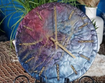 May include: A round, resin clock with a blue, purple, and pink marbled design. The clock face has gold-colored hour markers and hands. The clock is on a black metal stand.