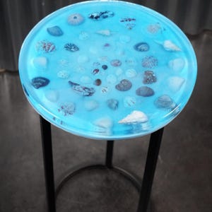 Pode incluir: Uma mesa lateral redonda com um tampo de resina azul claro com várias conchas embutidas. A mesa tem uma estrutura de metal preto e uma base circular. O tampo tem aproximadamente 46 cm de diâmetro.