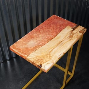 Puede incluir: Una mesa auxiliar rectangular que presenta una tabla de madera con borde natural unida a una sección de resina translúcida de color coral. El tablero se apoya sobre una estructura de metal dorado con una base en forma de C.