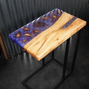 May include: A rectangular side table featuring a natural olive wood slab joined with a deep purple resin section. The resin contains embedded organic wood burl patterns. The tabletop is supported by a black metal frame with a C-shaped base design.