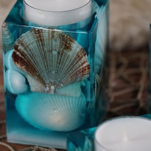 May include: Square teal resin candle holders with seashells and a white tea light candle inside. The candle holders have a beach theme, with a large scallop shell and smaller white shells embedded in the resin.