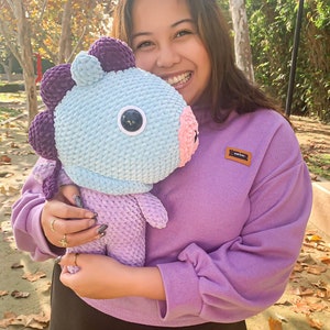 May include: A person is holding a crocheted plush toy that is light blue, purple, and pink. The toy has a large, round eye and a small, pink nose. The person is wearing a purple sweatshirt with a small brown patch that says "ARTIC" on it.