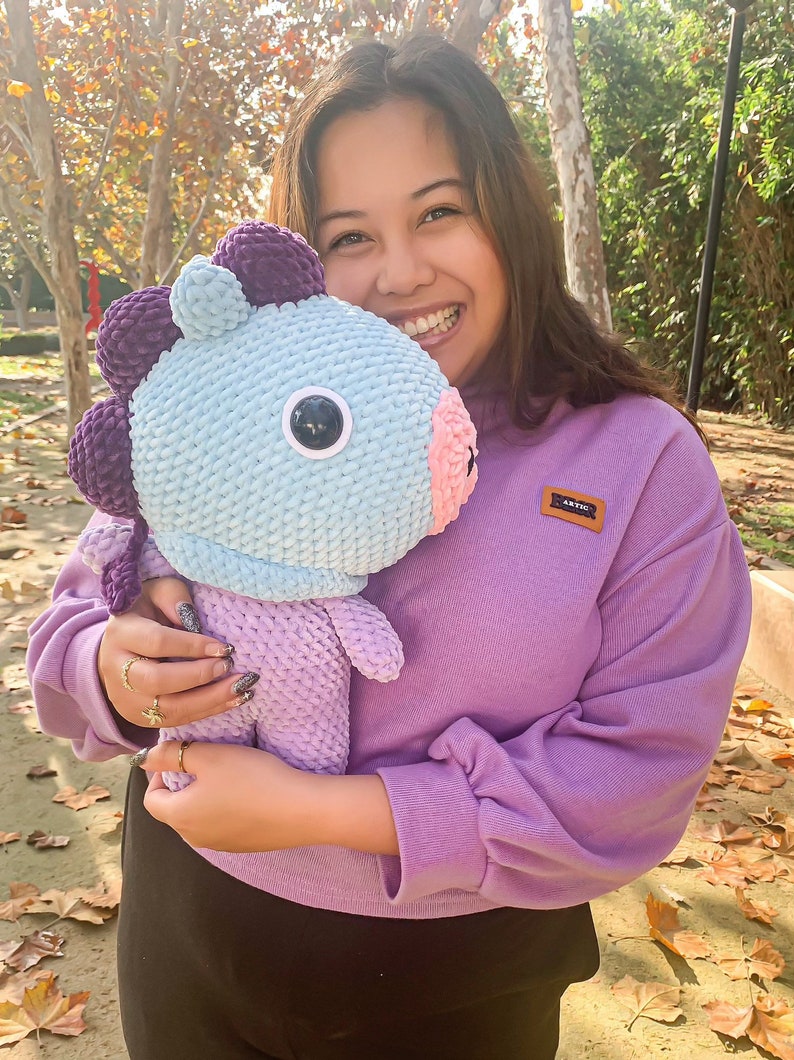 May include: A person is holding a crocheted plush toy that is light blue, purple, and pink. The toy has a large, round eye and a small, pink nose. The person is wearing a purple sweatshirt with a small brown patch that says "ARTIC" on it.