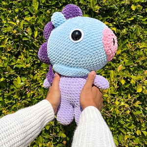 May include: A crocheted plush toy in the shape of a purple and blue horse with a pink nose and one large black eye. The toy is being held by a person wearing a white sweater.