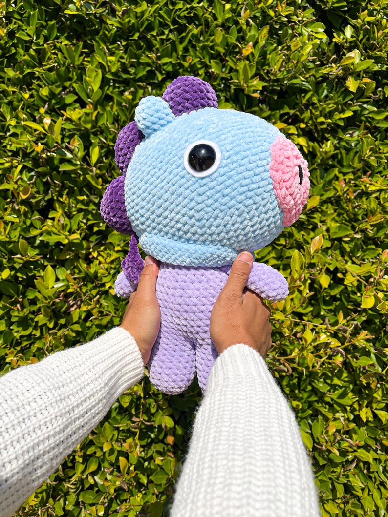 May include: A crocheted plush toy in the shape of a purple and blue horse with a pink nose and one large black eye. The toy is being held by a person wearing a white sweater.