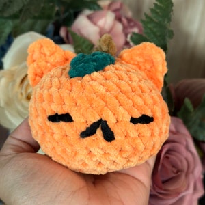 May include: A crocheted orange pumpkin with a green stem and black eyes and mouth. The pumpkin is held in a hand.