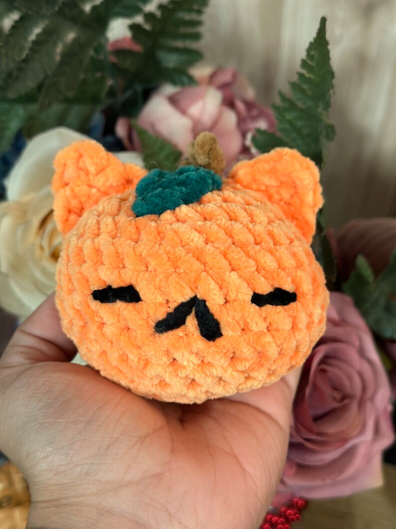 May include: A crocheted orange pumpkin with a green stem and black eyes and mouth. The pumpkin is held in a hand.