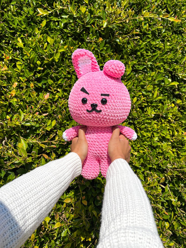 May include: A pink crocheted bunny plush toy with black eyes and a frowning expression. The toy is being held by hands wearing a white sweater.
