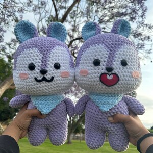 Puede incluir: Dos peluches de crochet con forma de animal de dibujos animados con pelaje morado y blanco, orejas azules y un babero azul. Los juguetes se toman de la mano y tienen caras sonrientes.