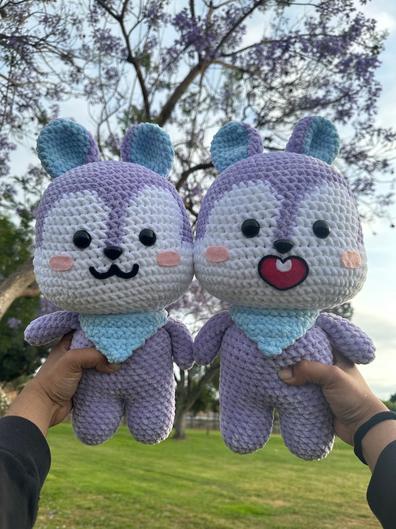 Puede incluir: Dos peluches de crochet con forma de animal de dibujos animados con pelaje morado y blanco, orejas azules y un babero azul. Los juguetes se toman de la mano y tienen caras sonrientes.