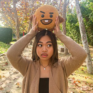 May include: A woman wearing a brown cardigan and a black top holds a brown plush toy with a cartoon face above her head. The toy has a frowning expression with a single tooth showing. The woman is standing in a park with fallen leaves on the ground.