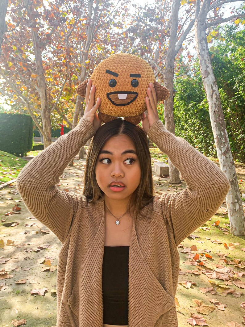 May include: A woman wearing a brown cardigan and a black top holds a brown plush toy with a cartoon face above her head. The toy has a frowning expression with a single tooth showing. The woman is standing in a park with fallen leaves on the ground.