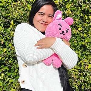 May include: A person wearing a white sweater with buttons is holding a pink crocheted bunny plush toy. The bunny has a black embroidered face with a frown and a small white nose.