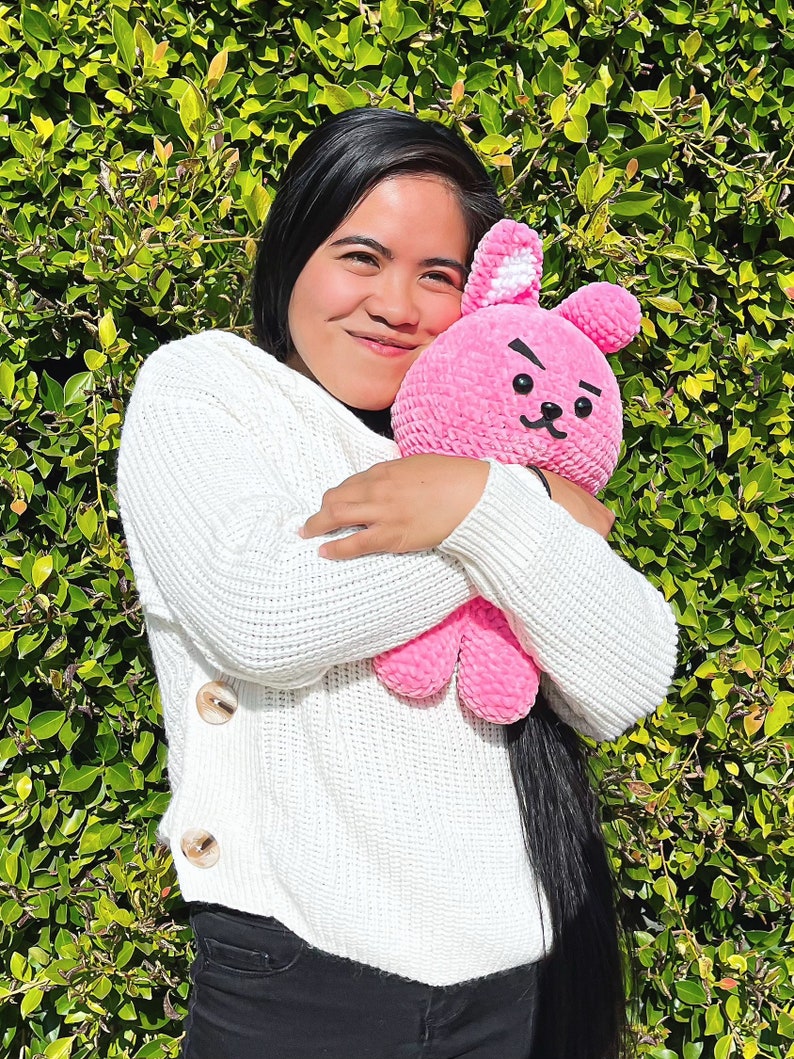 May include: A person wearing a white sweater with buttons is holding a pink crocheted bunny plush toy. The bunny has a black embroidered face with a frown and a small white nose.