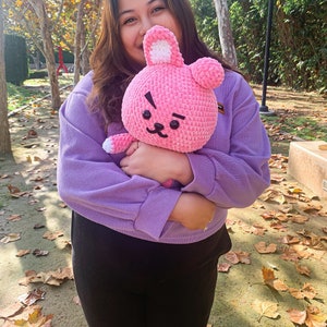 May include: A person wearing a purple sweater and black pants is holding a pink crocheted bunny plush toy. The toy has a black eye and a black nose.
