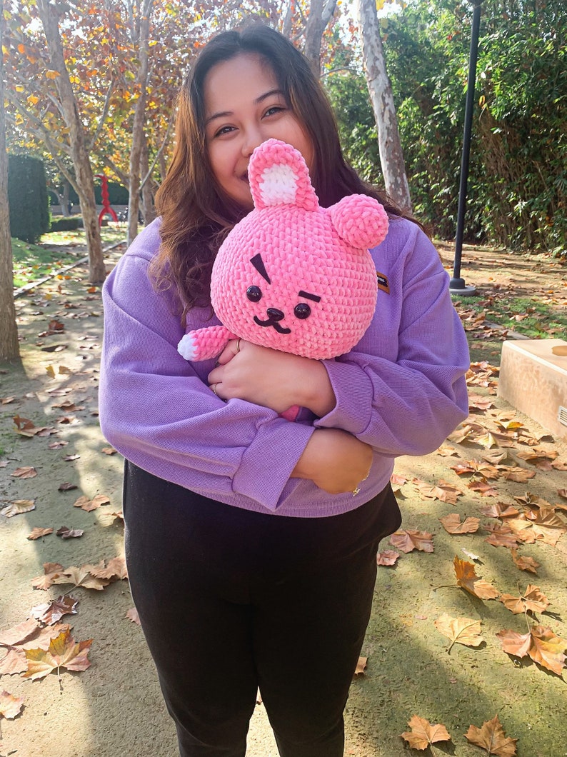 May include: A person wearing a purple sweater and black pants is holding a pink crocheted bunny plush toy. The toy has a black eye and a black nose.