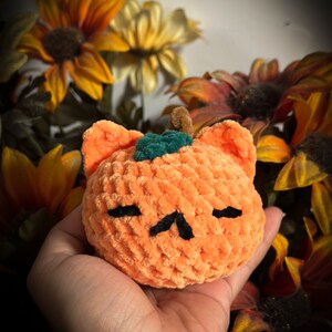 May include: A crocheted orange pumpkin with a green stem and black eyes. The pumpkin is held in a hand against a background of fall-colored flowers.
