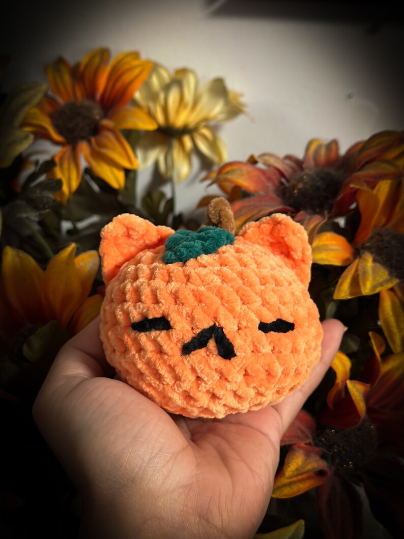 May include: A crocheted orange pumpkin with a green stem and black eyes. The pumpkin is held in a hand against a background of fall-colored flowers.