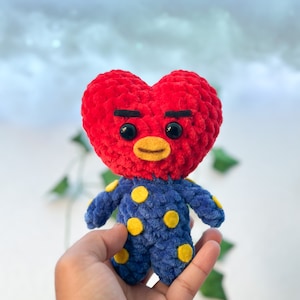 K&ouml;nnte beinhalten: Ein rotes, herzf&ouml;rmiges, geh&auml;keltes Pl&uuml;schtier mit einem blauen und gelben Tupfen-Outfit. Das Spielzeug hat schwarze Augenbrauen und gro&szlig;e schwarze Augen.