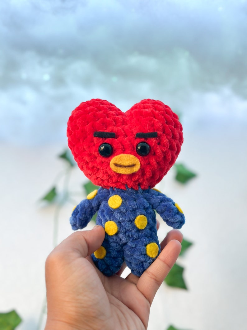 K&ouml;nnte beinhalten: Ein rotes, herzf&ouml;rmiges, geh&auml;keltes Pl&uuml;schtier mit einem blauen und gelben Tupfen-Outfit. Das Spielzeug hat schwarze Augenbrauen und gro&szlig;e schwarze Augen.