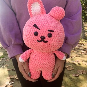 May include: A pink crocheted plush toy shaped like a rabbit with white ears and black eyes. The toy has a grumpy expression with a frown and a small mouth.