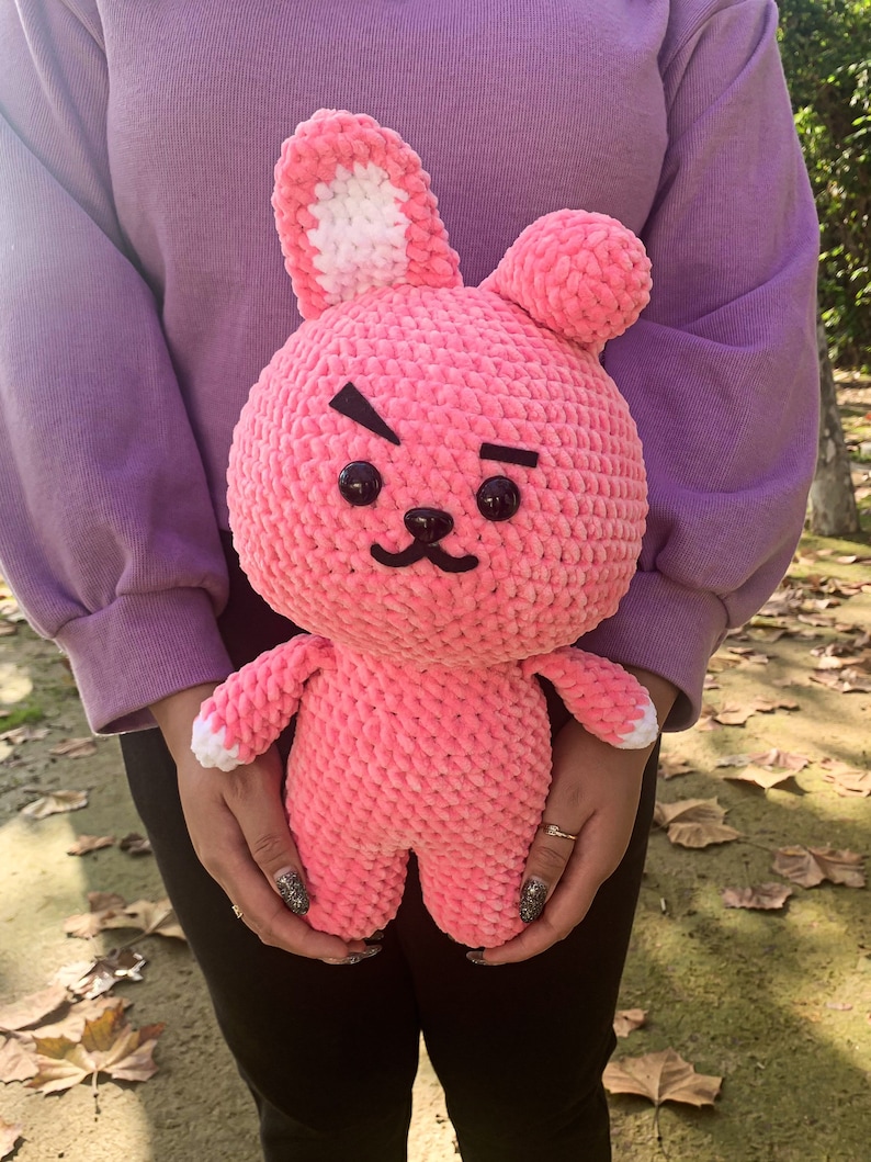May include: A pink crocheted plush toy shaped like a rabbit with white ears and black eyes. The toy has a grumpy expression with a frown and a small mouth.
