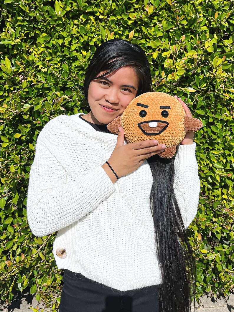 May include: A person wearing a white sweater is holding a brown plush toy with a smiling face. The toy has a large tooth and black eyes. The person has long black hair and is smiling.