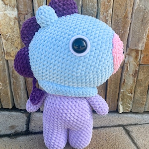 May include: A crocheted plush toy in the shape of a cartoon character. The toy is light blue with purple ears and a purple tail. It has a large, black eye and a pink nose.