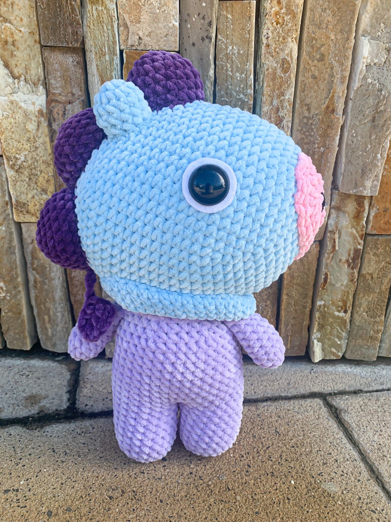 May include: A crocheted plush toy in the shape of a cartoon character. The toy is light blue with purple ears and a purple tail. It has a large, black eye and a pink nose.